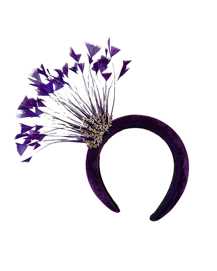 Still life picture of the Seraphina headband fascinator, wrapped in plush violet velvet. This sculptural piece bursts into movement with a spray of deep purple feathers and a gold-coloured metal fan featuring branch detailing on the right front side. Designed by Merve Bayindir.