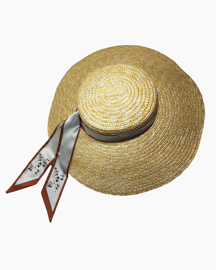 Natural woven straw hat with a wide brim, wrapped in a taupe and rust silk ribbon and finished with long scarf ties featuring delicate illustrated figures. Designed and made by Merve Bayindir Millinery.
