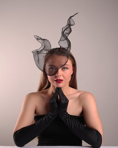 Model is showcasing the front view of the black crinoline, decorated with silver coloured star sequins, is wired and asymmetrically placed on a custom headband covering half of the face creating an illusion of a flying ribbon.