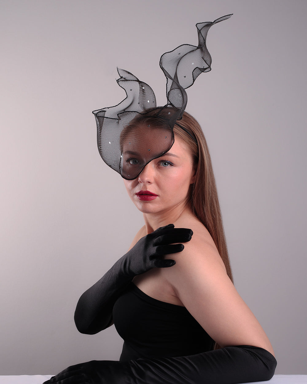 Model is showcasing the front left view of the black crinoline, decorated with silver coloured star sequins, is wired and asymmetrically placed on a custom headband covering half of the face creating an illusion of a flying ribbon.
