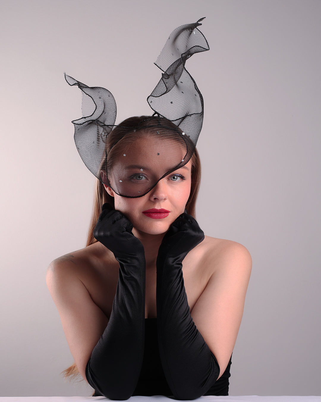 Model is showcasing the front right view of the black crinoline, decorated with silver coloured star sequins, is wired and asymmetrically placed on a custom headband covering half of the face creating an illusion of a flying ribbon.
