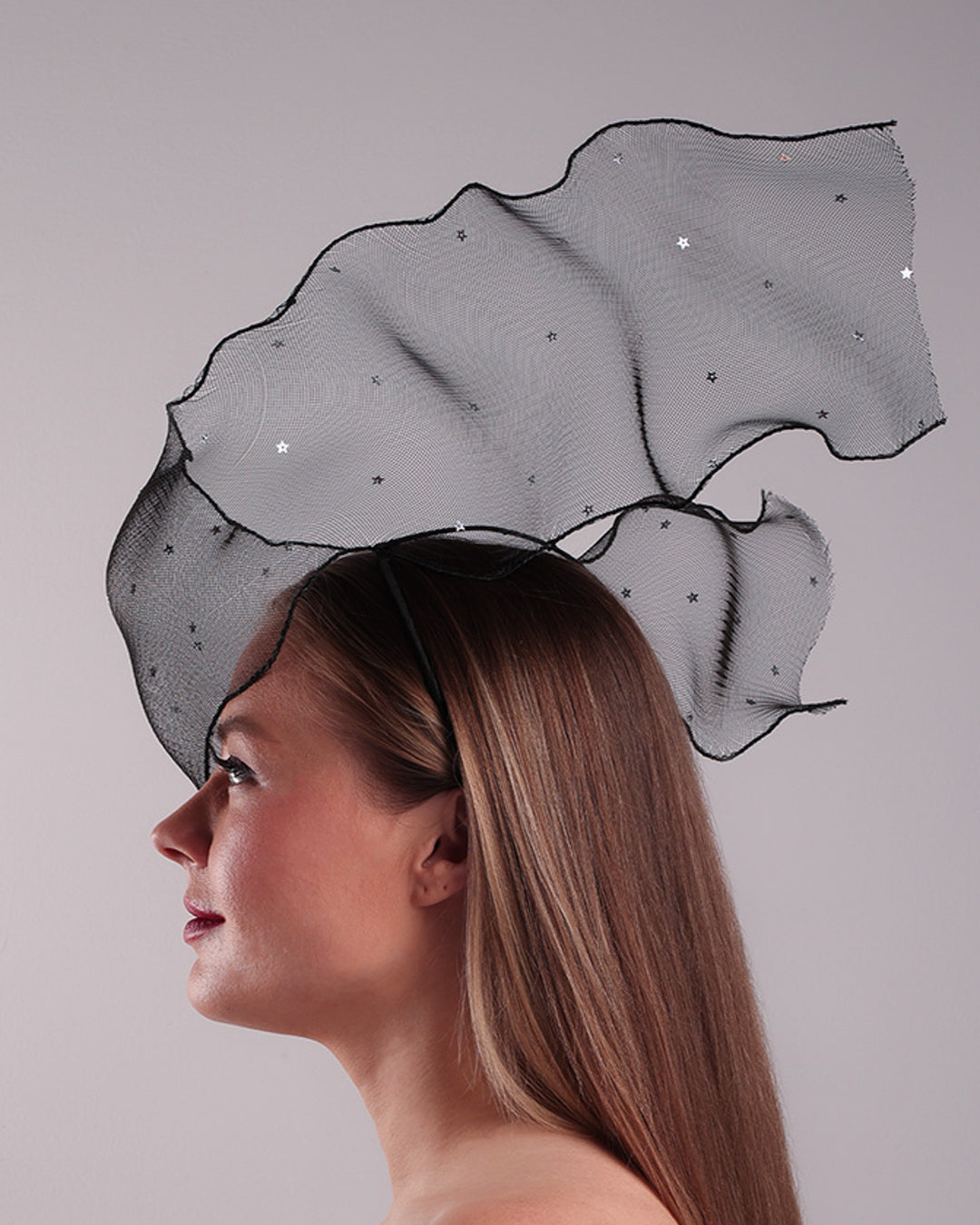 Model is showcasing the left side of the black crinoline, decorated with silver coloured star sequins, is wired and asymmetrically placed on a custom headband covering half of the face creating an illusion of a flying ribbon.