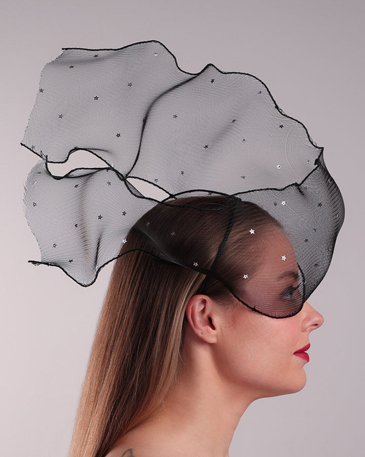 Model is showcasing the right side of the black crinoline, decorated with silver coloured star sequins, is wired and asymmetrically placed on a custom headband covering half of the face creating an illusion of a flying ribbon.