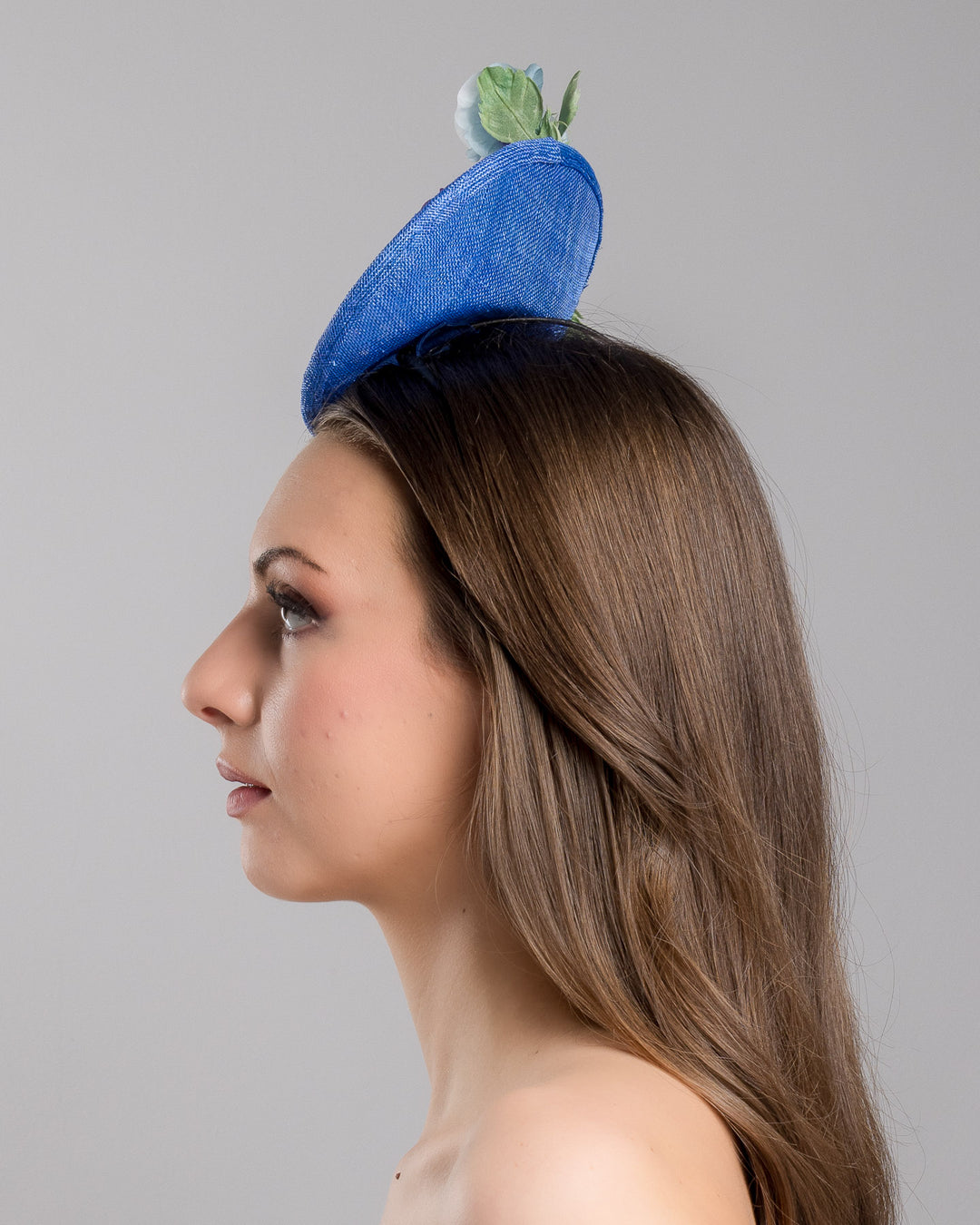 The model is showcasing the left side close up of the deep blue saucer style mini fascinator, decorate with different coloured violet flowers. Designed and made by Merve Bayindir Millinery.

