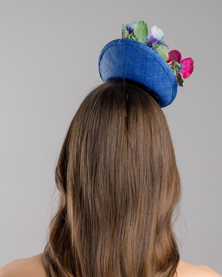 The model is showcasing the back side close up of the deep blue saucer style mini fascinator, decorate with different coloured violet flowers. Designed and made by Merve Bayindir Millinery.
