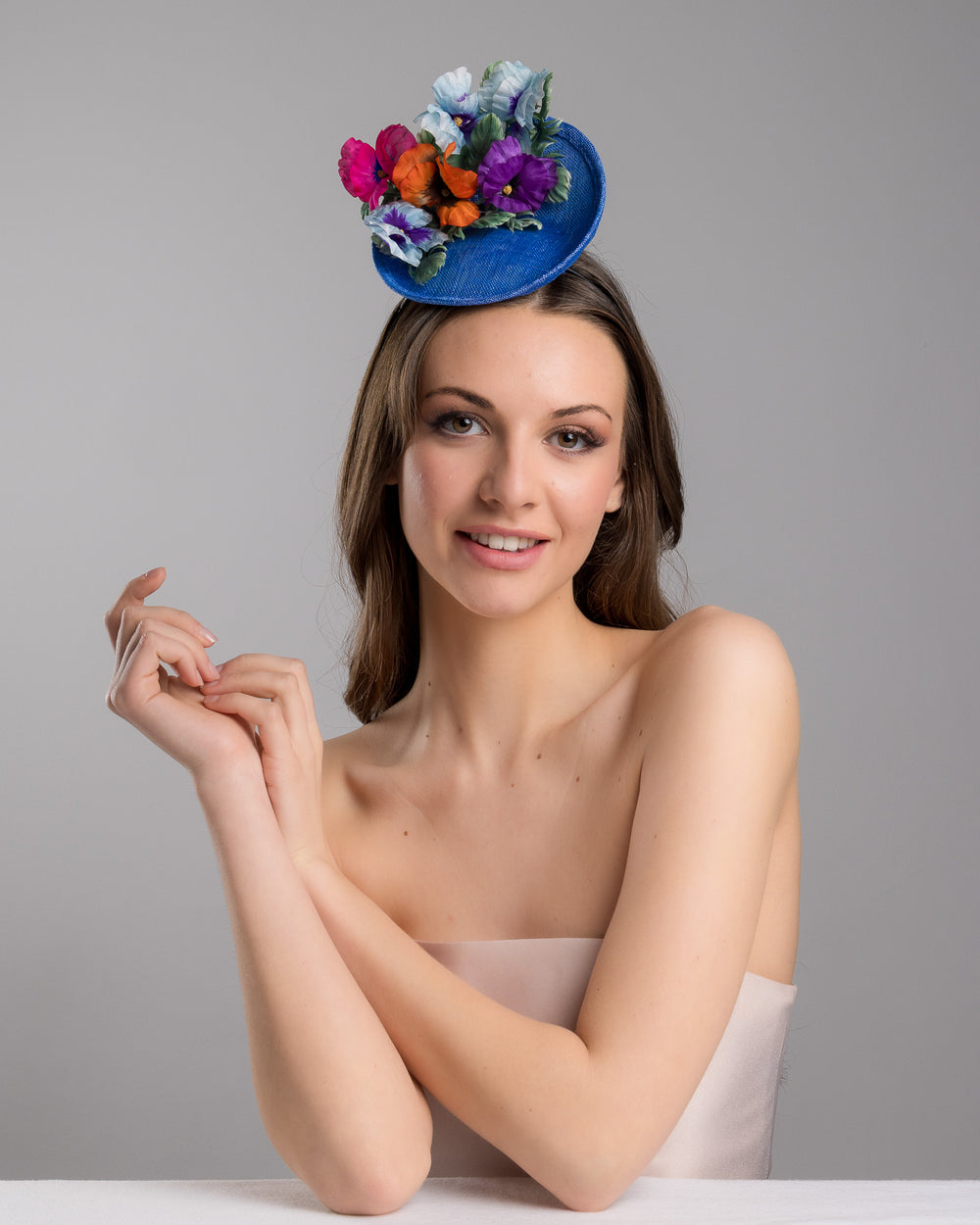 The female model is posing, wearing Shawn, the deep blue saucer style mini fascinator, decorate with different coloured violet flowers. Designed and made by Merve Bayindir Millinery.
