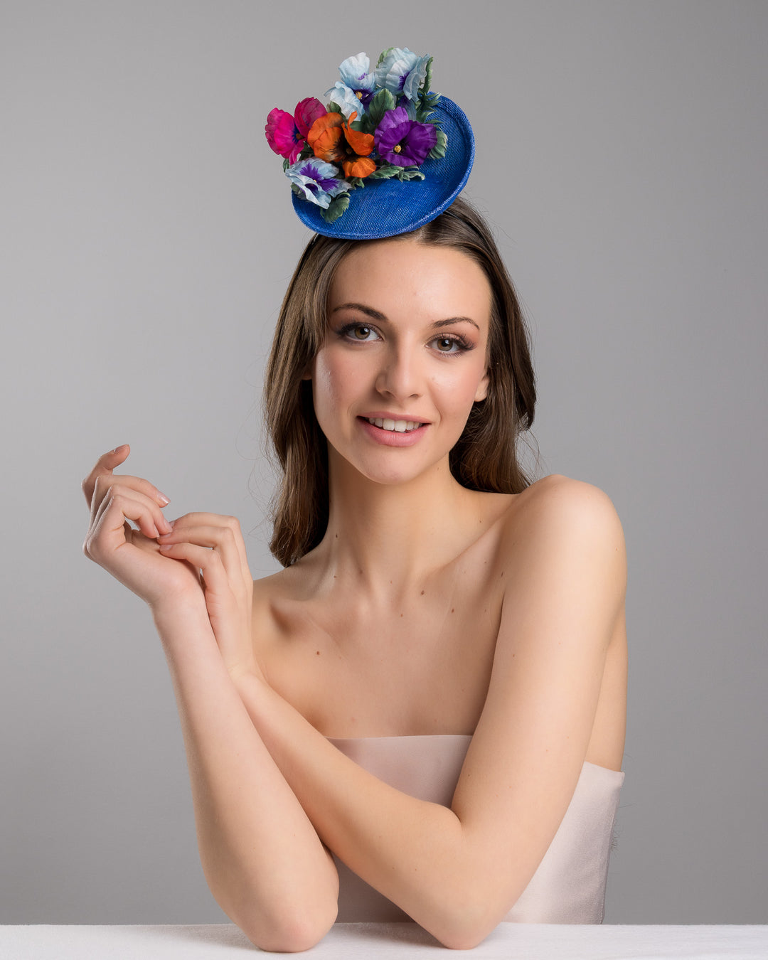 The female model is posing, wearing Shawn, the deep blue saucer style mini fascinator, decorate with different coloured violet flowers. Designed and made by Merve Bayindir Millinery.
