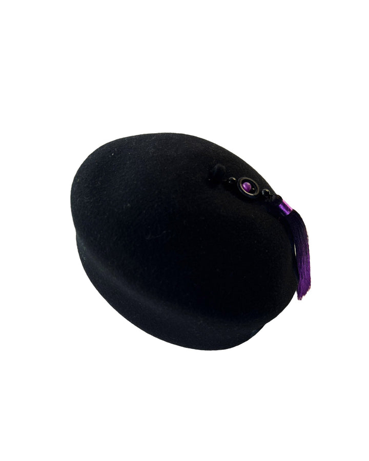 Still life picture of the Sienna Hat, shaped from structured black wool felt, it features a bold, angled crown and a striking violet tassel that adds a touch of colour and movement on the right side. Designed by Merve Bayindir.