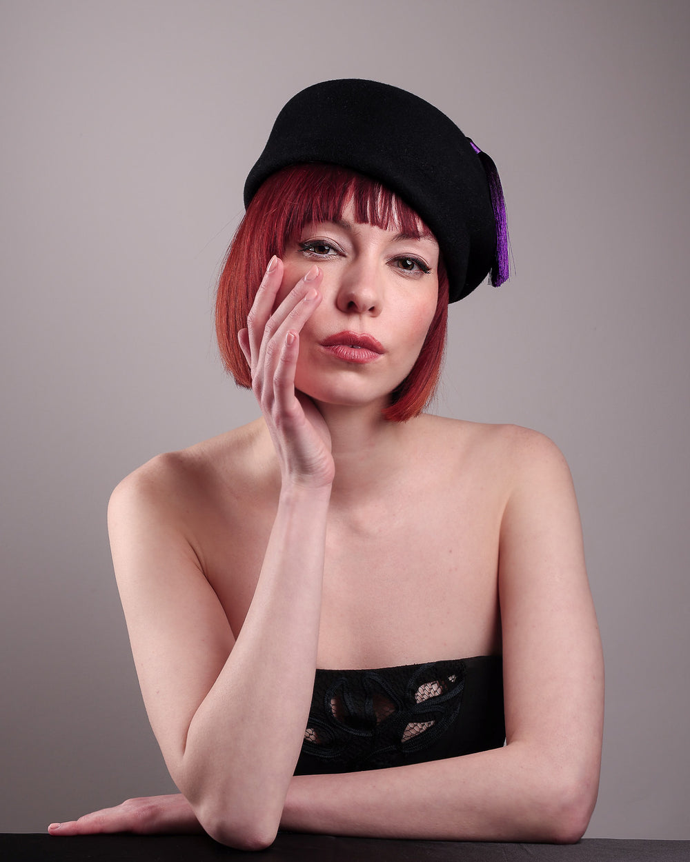The model is showcasing the front of the Sien Hat, shaped from structured black wool felt, it features a bold, angled crown and a striking violet tassel that adds a touch of colour and movement on the right side. Designed by Merve Bayindir.