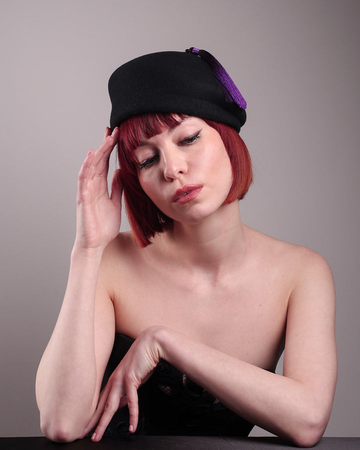 The model is showcasing the front below anfle of the Sien Hat, shaped from structured black wool felt, it features a bold, angled crown and a striking violet tassel that adds a touch of colour and movement on the right side. Designed by Merve Bayindir.
