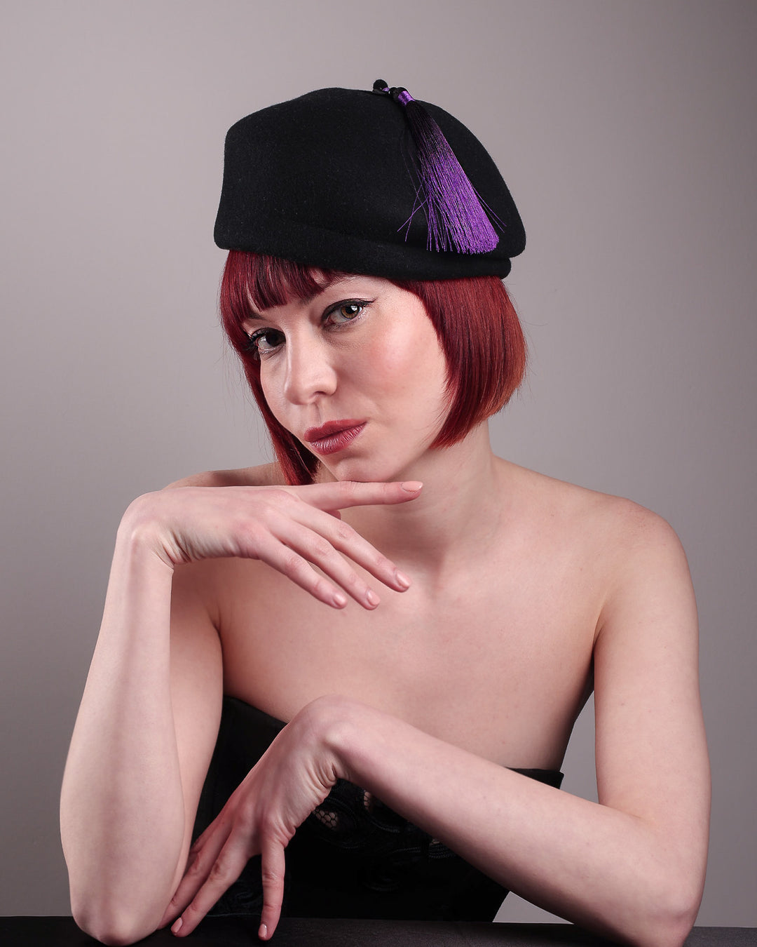 The model is showcasing the front right side of the Sien Hat, shaped from structured black wool felt, it features a bold, angled crown and a striking violet tassel that adds a touch of colour and movement on the right side. Designed by Merve Bayindir.