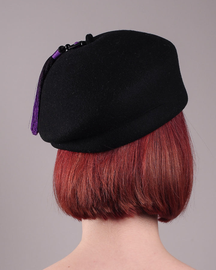 The model is showcasing the back of the Sien Hat, shaped from structured black wool felt, it features a bold, angled crown and a striking violet tassel that adds a touch of colour and movement on the right side. Designed by Merve Bayindir.