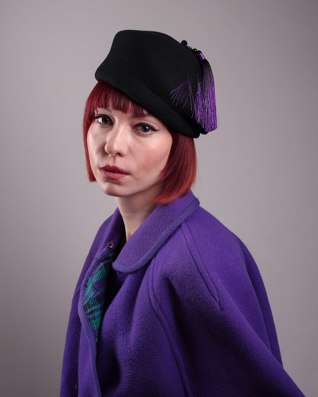 The model is showcasing the styled picture of the Sien Hat, shaped from structured black wool felt, it features a bold, angled crown and a striking violet tassel that adds a touch of colour and movement on the right side, matched with a purple felt coat. Designed by Merve Bayindir.