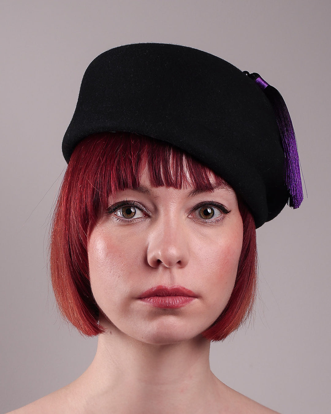 The model is showcasing the front close up of the Sien Hat, shaped from structured black wool felt, it features a bold, angled crown and a striking violet tassel that adds a touch of colour and movement on the right side. Designed by Merve Bayindir.