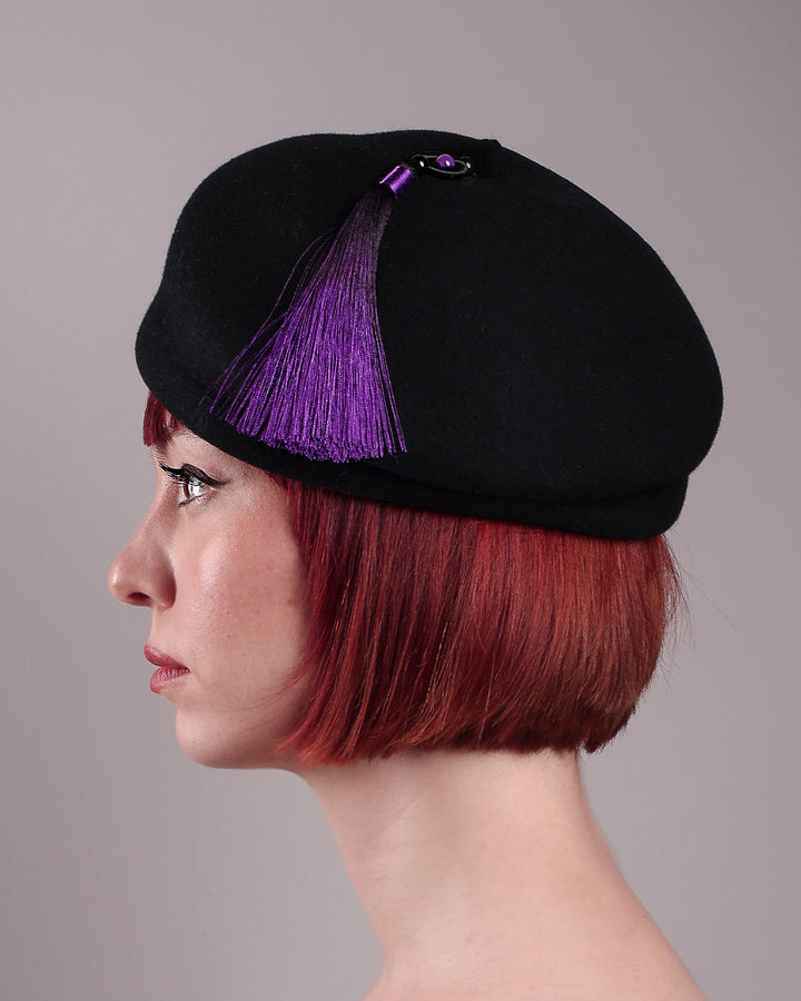 The model is showcasing the front close up of the Sien Hat, shaped from structured black wool felt, it features a bold, angled crown and a striking violet tassel that adds a touch of colour and movement on the right side. Designed by Merve Bayindir.