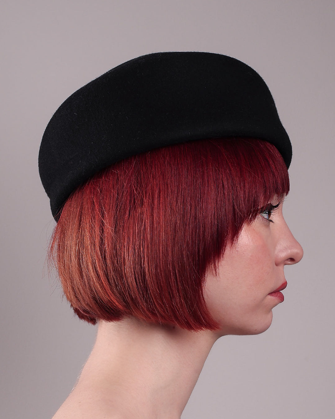 The model is showcasing the right side close up of the Sien Hat, shaped from structured black wool felt, it features a bold, angled crown and a striking violet tassel that adds a touch of colour and movement on the right side. Designed by Merve Bayindir.