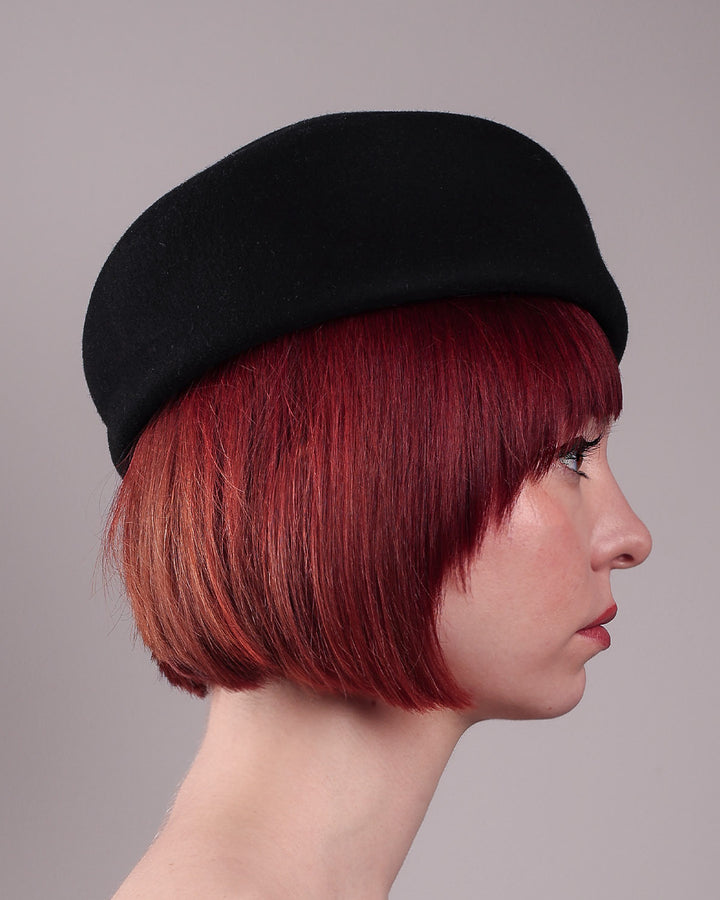 The model is showcasing the right side close up of the Sien Hat, shaped from structured black wool felt, it features a bold, angled crown and a striking violet tassel that adds a touch of colour and movement on the right side. Designed by Merve Bayindir.