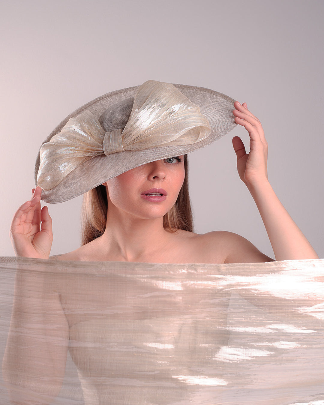 Model is showcasing the front of a grey asymetric wide-brim hat decorated with striking oversized bow with silver metallic details. The bow matches the shape of the asymety of the hat. Worn to tilted to the right side of the head, fitted with a wire and elastic.