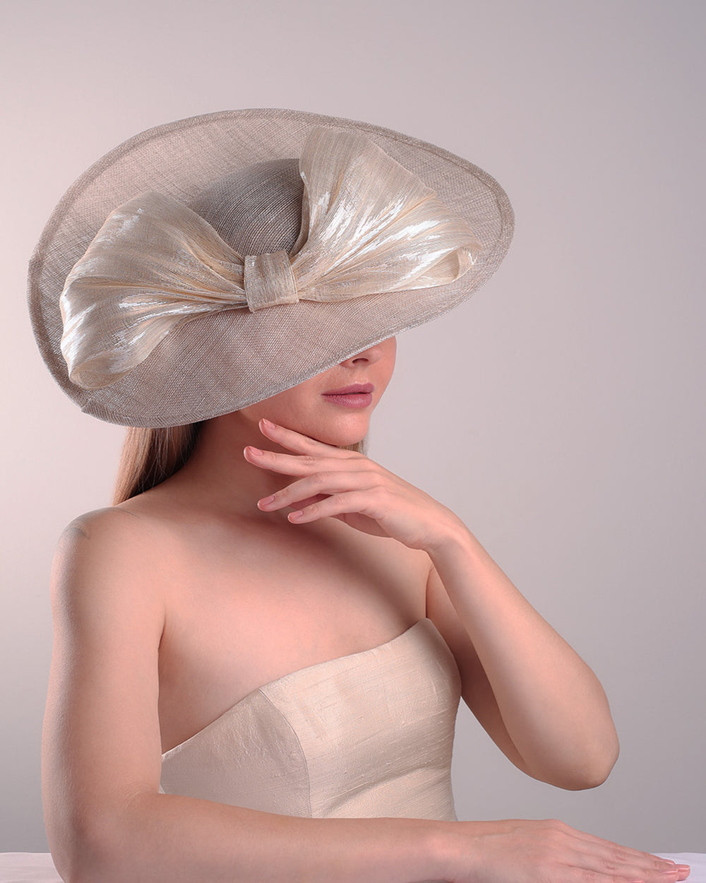 Model is showcasing the right top front side of a grey asymetric wide-brim hat decorated with striking oversized bow with silver metallic details. The bow matches the shape of the asymety of the hat. Worn to tilted to the right side of the head, fitted with a wire and elastic.