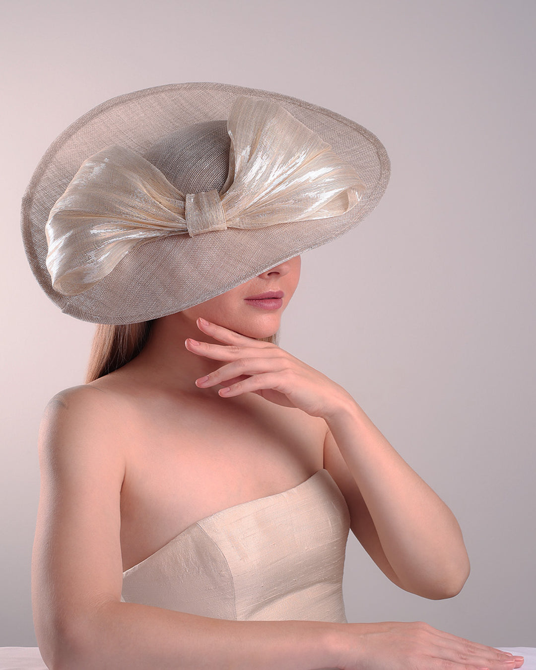 Model is showcasing the right top front side of a grey asymetric wide-brim hat decorated with striking oversized bow with silver metallic details. The bow matches the shape of the asymety of the hat. Worn to tilted to the right side of the head, fitted with a wire and elastic.