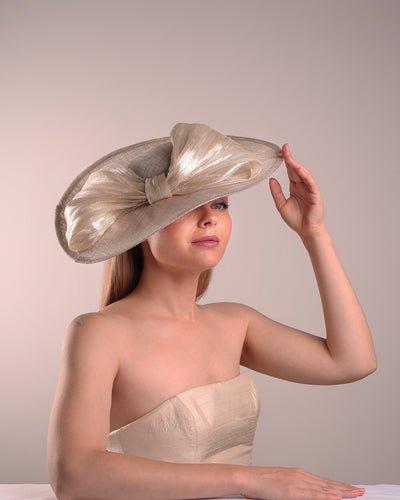 Model is showcasing the right front side of a grey asymetric wide-brim hat decorated with striking oversized bow with silver metallic details. The bow matches the shape of the asymety of the hat. Worn to tilted to the right side of the head, fitted with a wire and elastic.