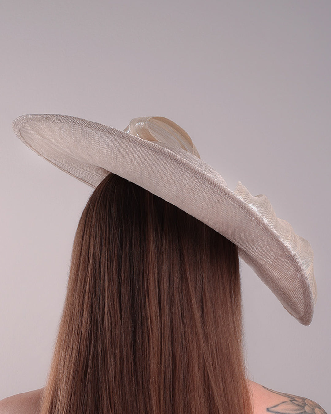 Model is showcasing the back of a grey asymetric wide-brim hat decorated with striking oversized bow with silver metallic details. The bow matches the shape of the asymety of the hat. Worn to tilted to the right side of the head, fitted with a wire and elastic.