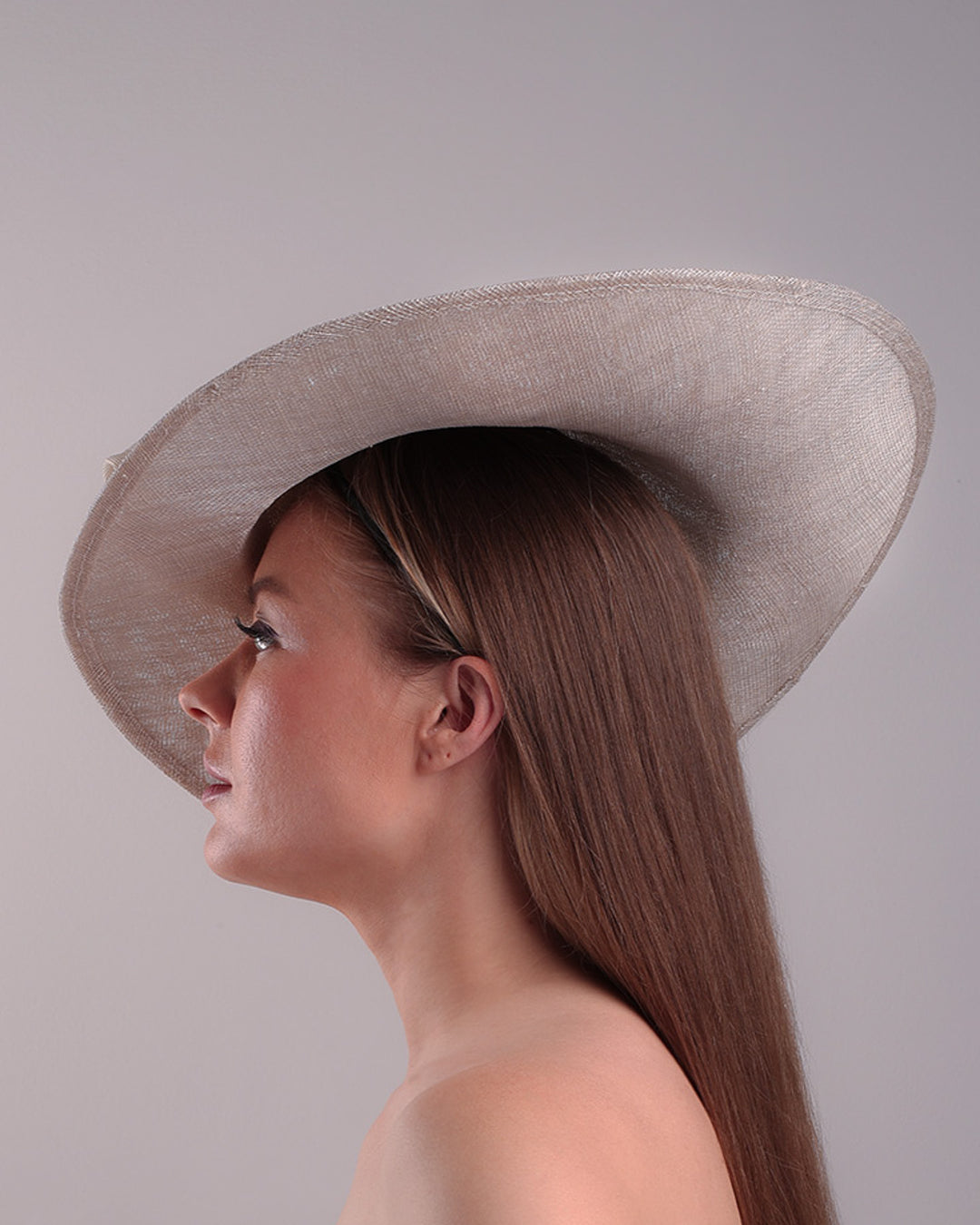 Model is showcasing the left side of a grey asymetric wide-brim hat decorated with striking oversized bow with silver metallic details. The bow matches the shape of the asymety of the hat. Worn to tilted to the right side of the head, fitted with a wire and elastic.