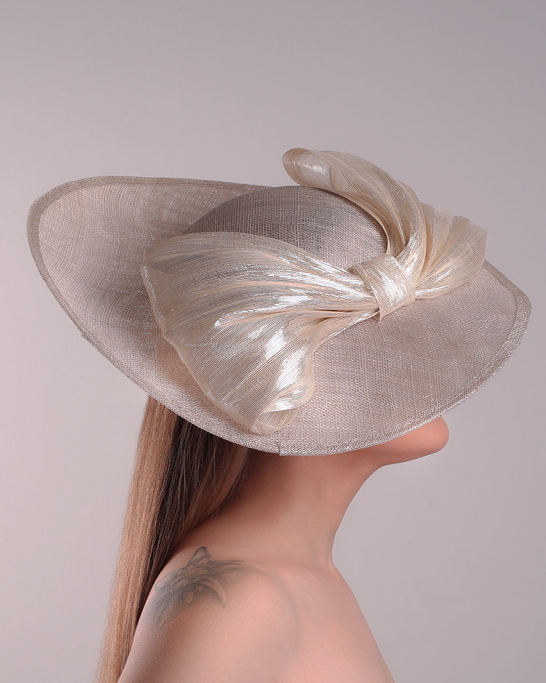 Model is showcasing the right side of a grey asymetric wide-brim hat decorated with striking oversized bow with silver metallic details. The bow matches the shape of the asymety of the hat. Worn to tilted to the right side of the head, fitted with a wire and elastic.