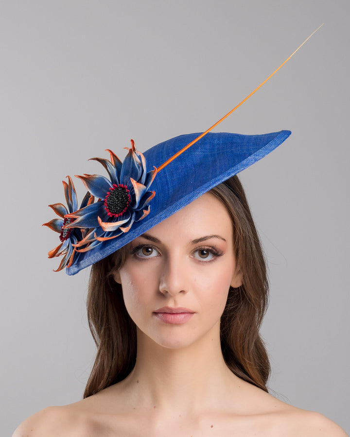 The model is showcasing the front side close up of the deep blue disk style wide brim hat, decorated with blue sunflower style flowers with orange edges, its embellished with an orange coloured ostrich quill. Designed and made by Merve Bayindir Millinery.