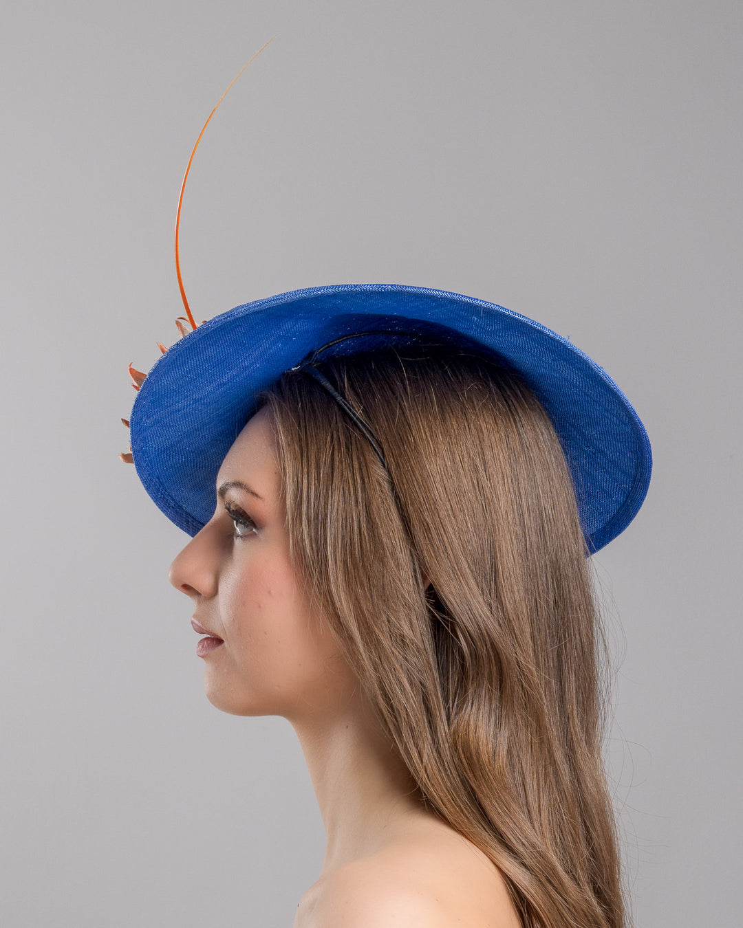 The model is showcasing the left side close up of the deep blue disk style wide brim hat, decorated with blue sunflower style flowers with orange edges, its embellished with an orange coloured ostrich quill. Designed and made by Merve Bayindir Millinery.
