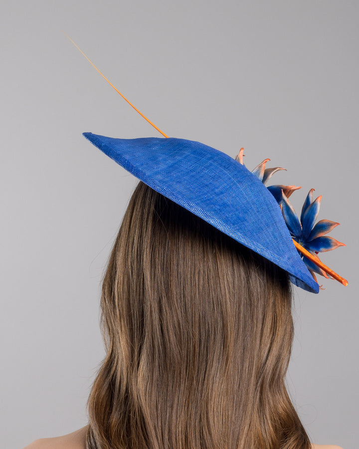The model is showcasing the back side close up of the deep blue disk style wide brim hat, decorated with blue sunflower style flowers with orange edges, its embellished with an orange coloured ostrich quill. Designed and made by Merve Bayindir Millinery.