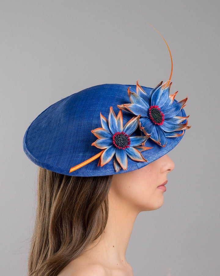 The model is showcasing the right side close up of the deep blue disk style wide brim hat, decorated with blue sunflower style flowers with orange edges, its embellished with an orange coloured ostrich quill. Designed and made by Merve Bayindir Millinery.

