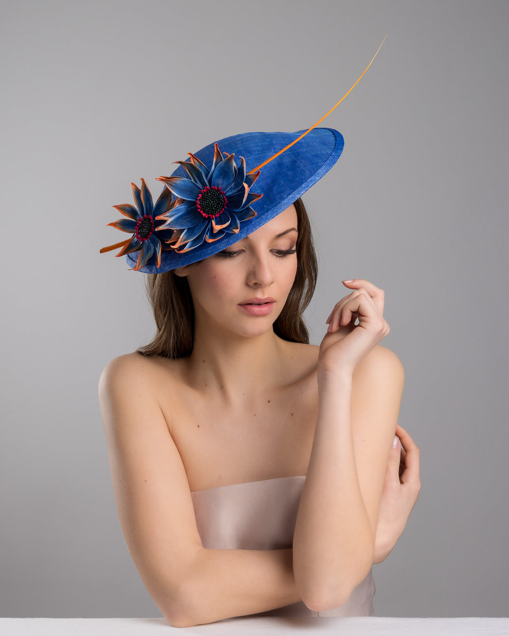 The female model is posing, wearing Sol,  the deep blue disk style wide brim hat, decorated with blue sunflower style flowers with orange edges, its embellished with an orange coloured ostrich quill. Designed and made by Merve Bayindir Millinery.