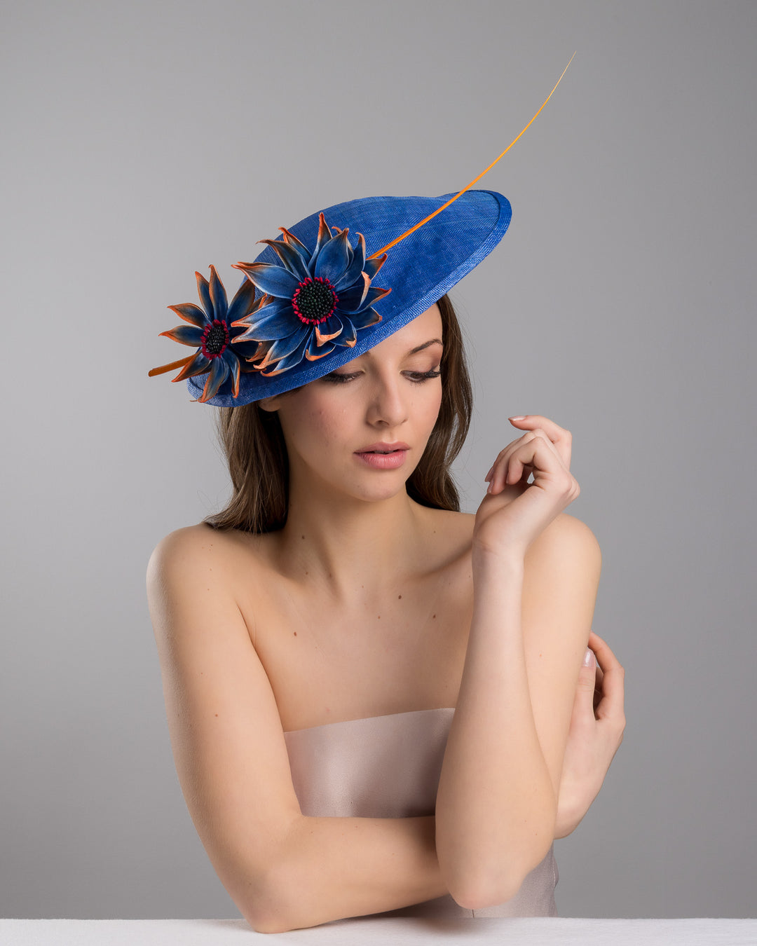 The female model is posing, wearing Sol,  the deep blue disk style wide brim hat, decorated with blue sunflower style flowers with orange edges, its embellished with an orange coloured ostrich quill. Designed and made by Merve Bayindir Millinery.