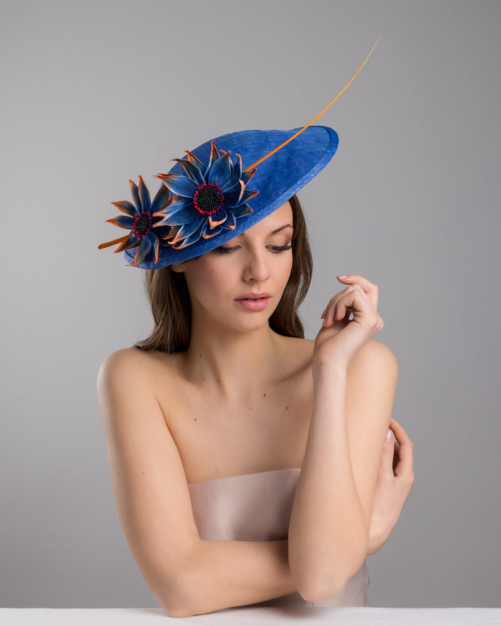 The female model is posing, wearing Sol,  the deep blue disk style wide brim hat, decorated with blue sunflower style flowers with orange edges, its embellished with an orange coloured ostrich quill. Designed and made by Merve Bayindir Millinery.