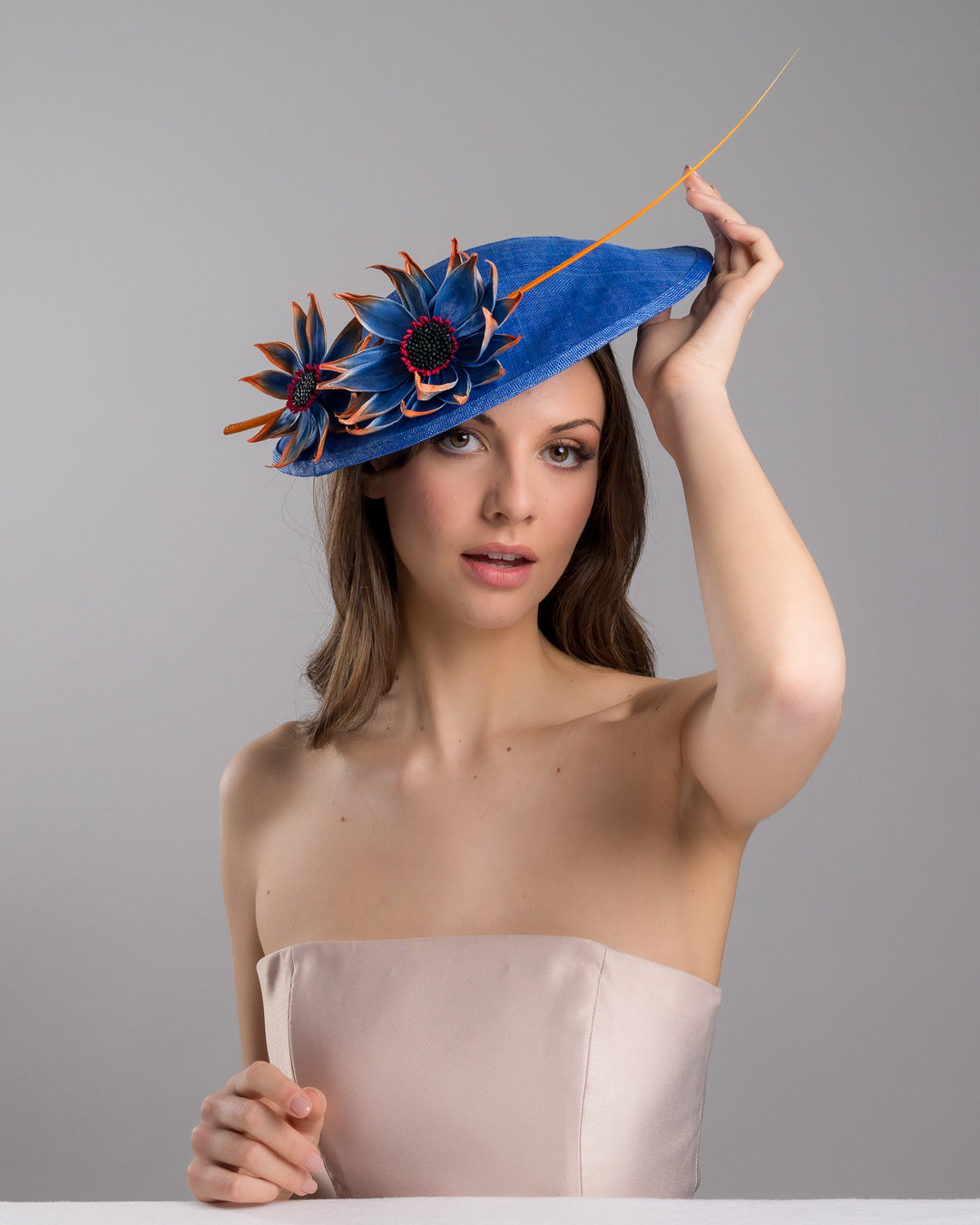 The female model is posing, wearing Sol,  the deep blue disk style wide brim hat, decorated with blue sunflower style flowers with orange edges, its embellished with an orange coloured ostrich quill. Designed and made by Merve Bayindir Millinery.