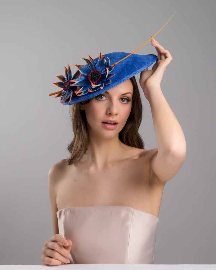 The female model is posing, wearing Sol,  the deep blue disk style wide brim hat, decorated with blue sunflower style flowers with orange edges, its embellished with an orange coloured ostrich quill. Designed and made by Merve Bayindir Millinery.