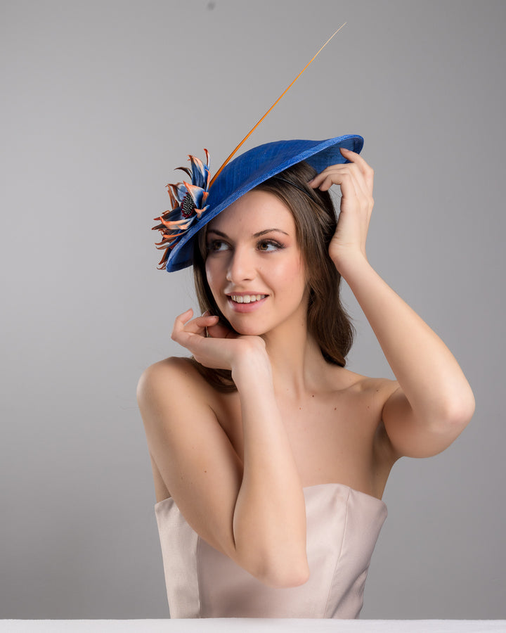 The female model is posing, wearing Sol,  the deep blue disk style wide brim hat, decorated with blue sunflower style flowers with orange edges, its embellished with an orange coloured ostrich quill. Designed and made by Merve Bayindir Millinery.