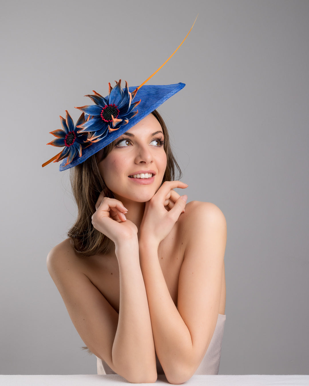The female model is posing, wearing Sol,  the deep blue disk style wide brim hat, decorated with blue sunflower style flowers with orange edges, its embellished with an orange coloured ostrich quill. Designed and made by Merve Bayindir Millinery.