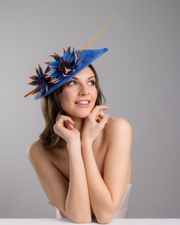 The female model is posing, wearing Sol,  the deep blue disk style wide brim hat, decorated with blue sunflower style flowers with orange edges, its embellished with an orange coloured ostrich quill. Designed and made by Merve Bayindir Millinery.