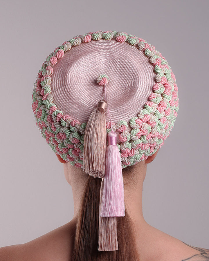 Model is presenting the back of a parasisal base pink pillbox hat that is decorated with pink, green and beige mix bead clusters,adorned with layered pink and beige tassels on the top.