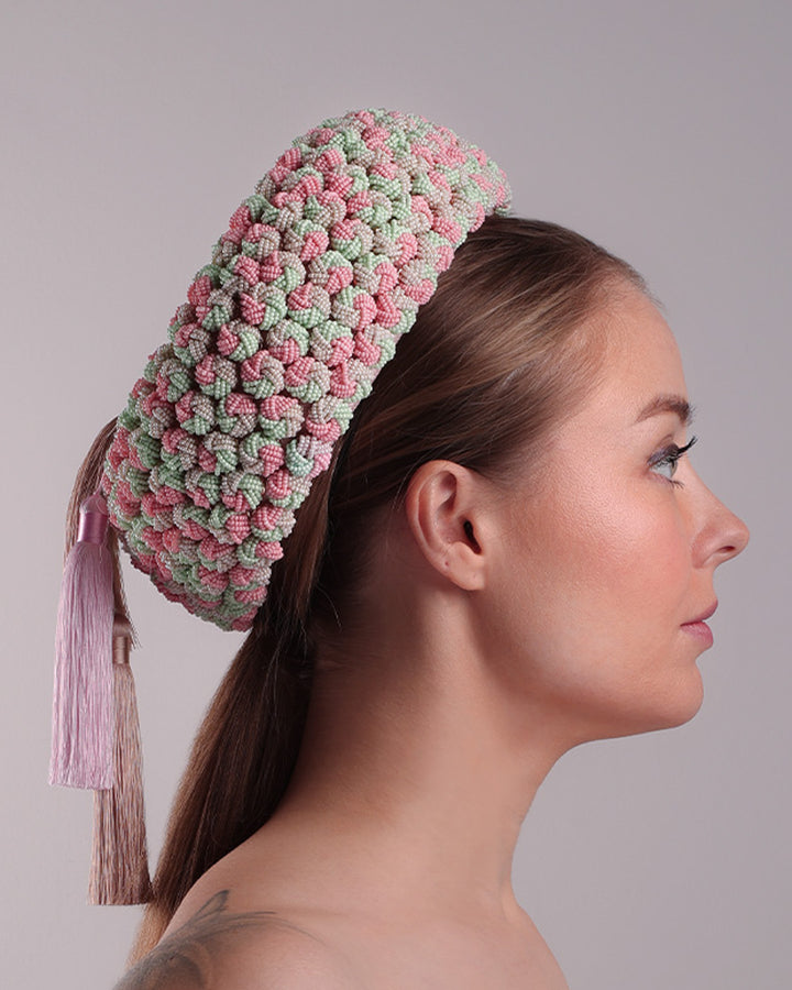 Model is presenting the right side of a parasisal base pink pillbox hat that is decorated with pink, green and beige mix bead clusters,adorned with layered pink and beige tassels on the top.