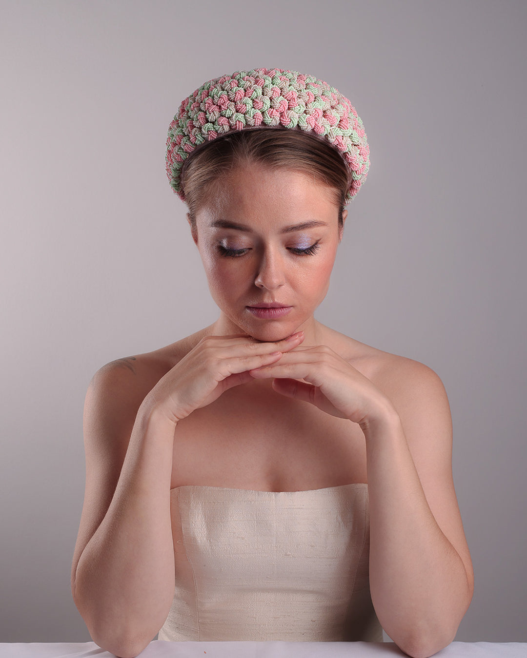 Model is presenting the fornt of a parasisal base pink pillbox hat that is decorated with pink, green and beige mix bead clusters,adorned with layered pink and beige tassels on the top.