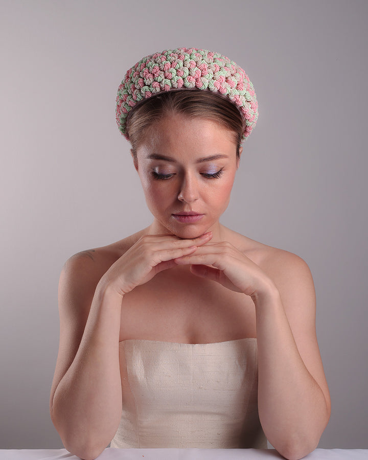 Model is presenting the fornt of a parasisal base pink pillbox hat that is decorated with pink, green and beige mix bead clusters,adorned with layered pink and beige tassels on the top.