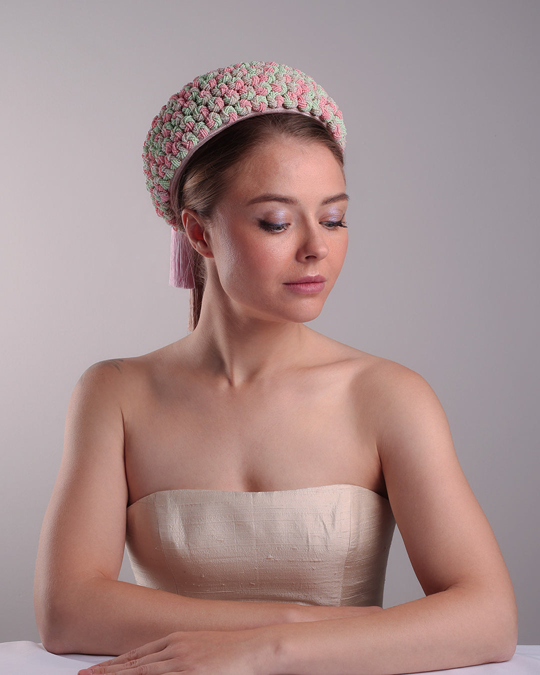 Model is presenting a parasisal base pink pillbox hat that is decorated with pink, green and beige mix bead clusters,adorned with layered pink and beige tassels on the top.