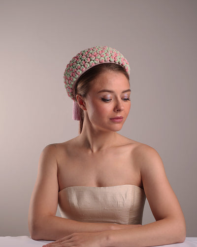 Model is presenting a parasisal base pink pillbox hat that is decorated with pink, green and beige mix bead clusters,adorned with layered pink and beige tassels on the top.