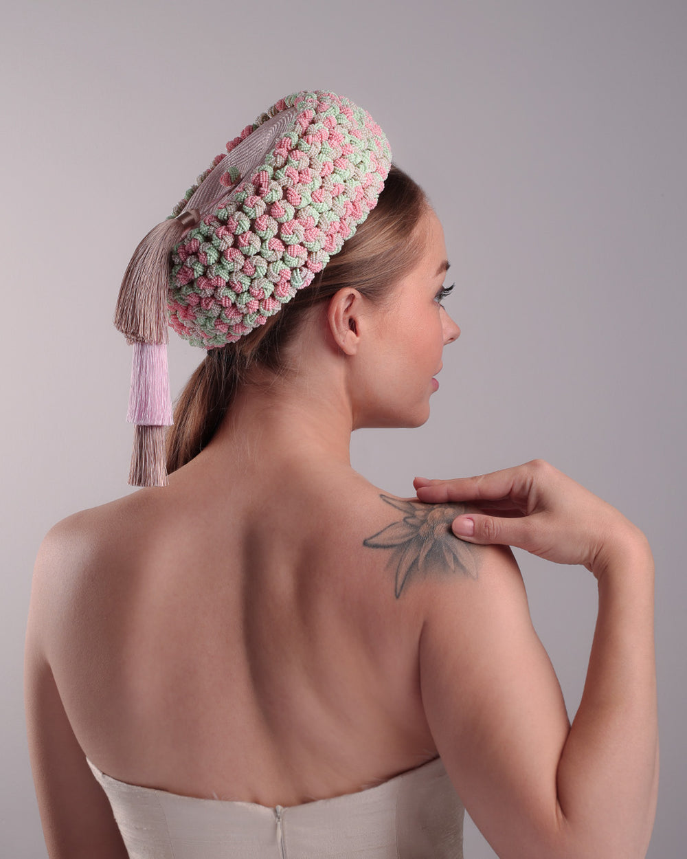 Model is presenting the right back and front angle of  parasisal base pink pillbox hat that is decorated with pink, green and beige mix bead clusters,adorned with layered pink and beige tassels on the top.