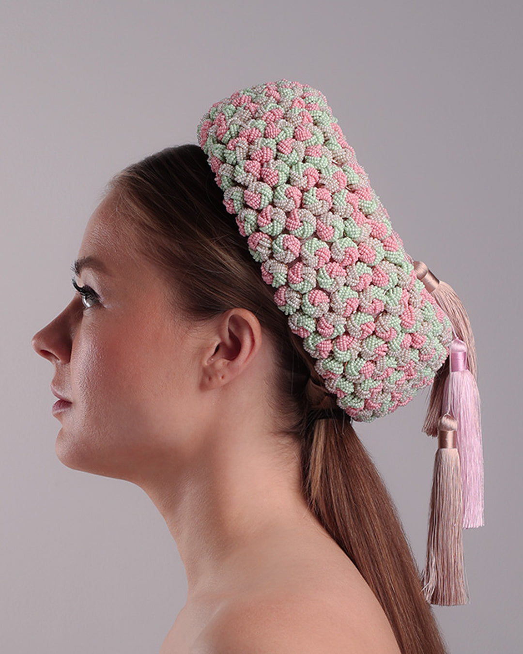 Model is presenting the left side of a parasisal base pink pillbox hat that is decorated with pink, green and beige mix bead clusters,adorned with layered pink and beige tassels on the top.