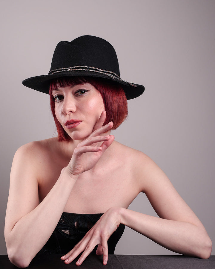 The model showcases the left front of the Stella fedora crafted in deep black felt, the silhouette is elevated by a subtle metallic crystal band featuring strands of crystal embellished thread that catch the light with every tilt of the head. The tied ends fall just off-centre on the left side. Designed by Merve Bayindir.