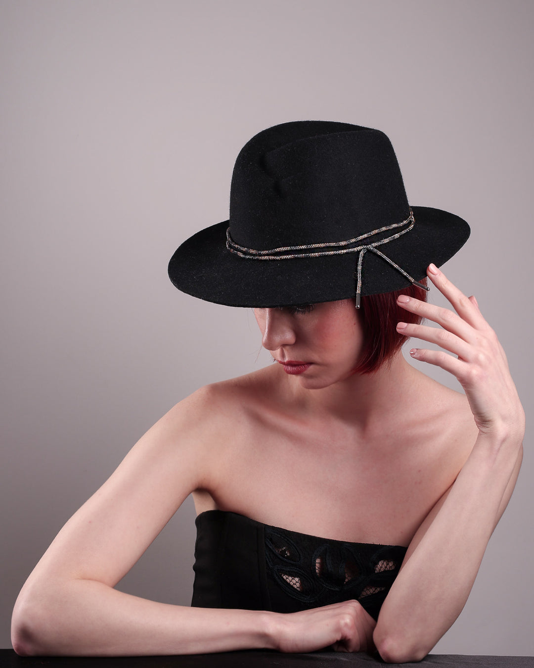 The model showcases the left high angle of the Stella fedora crafted in deep black felt, the silhouette is elevated by a subtle metallic crystal band featuring strands of crystal embellished thread that catch the light with every tilt of the head. The tied ends fall just off-centre on the left side. Designed by Merve Bayindir.